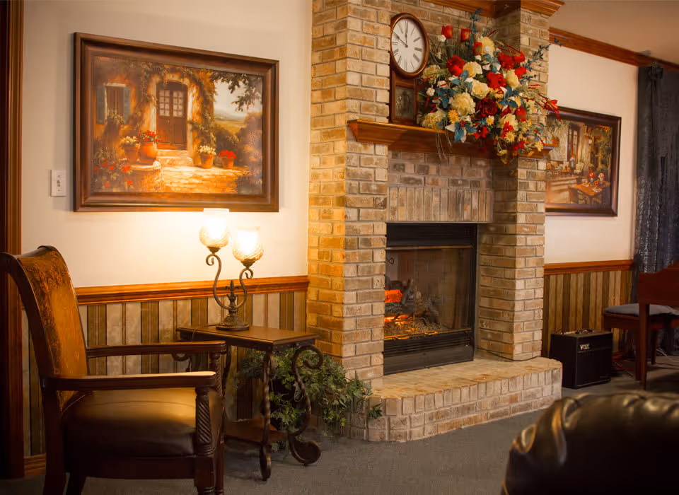 Cozy living room area with a brick fireplace, armchairs, framed paintings, and a side table with a lamp and floral mantel display.