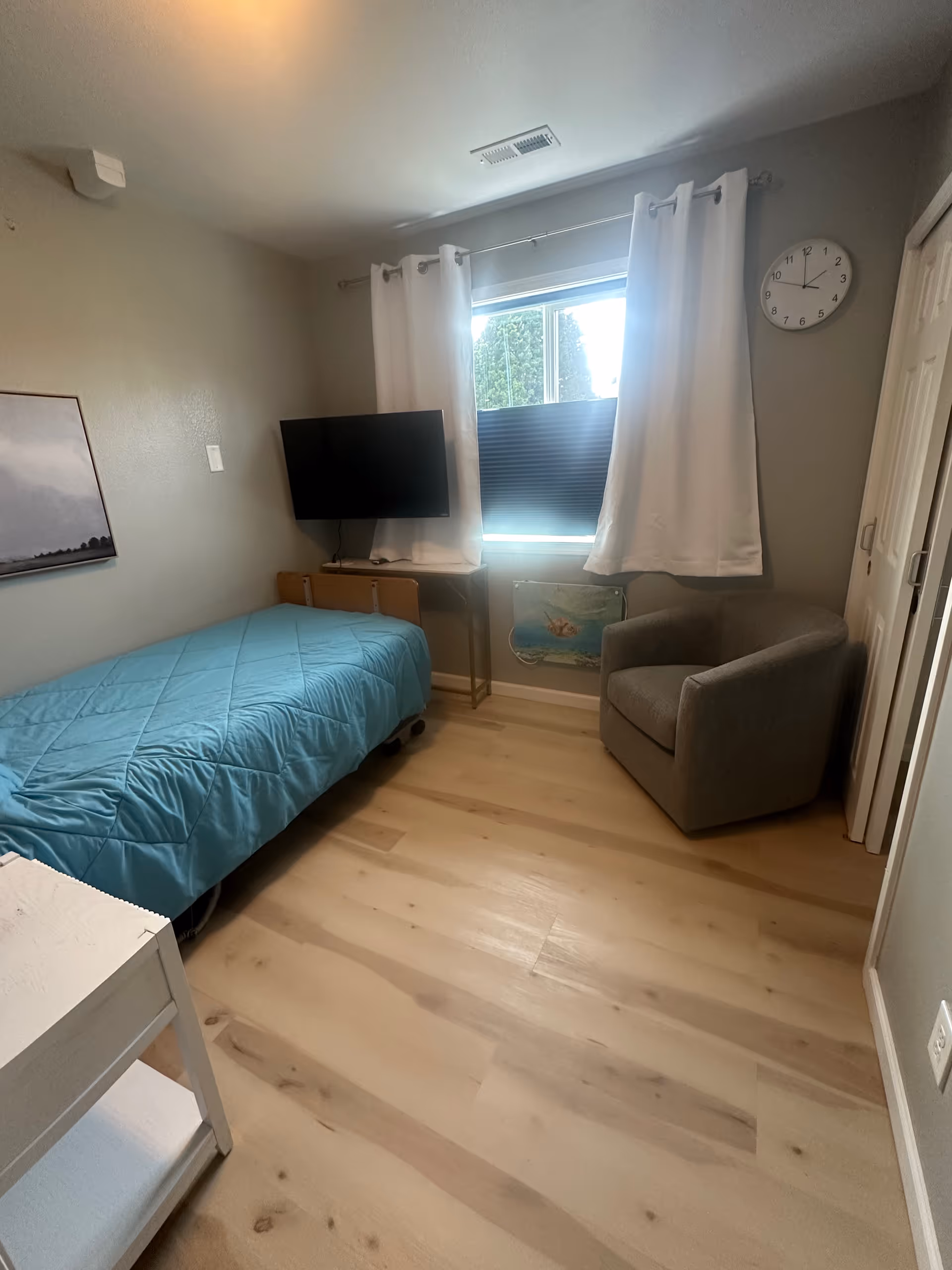 Small bedroom with blue bedspread, window, desk, TV, clock, and gray chair.