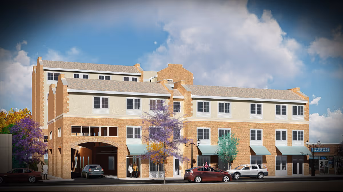 Three-story brick senior living building with storefronts and parked cars along a busy street.