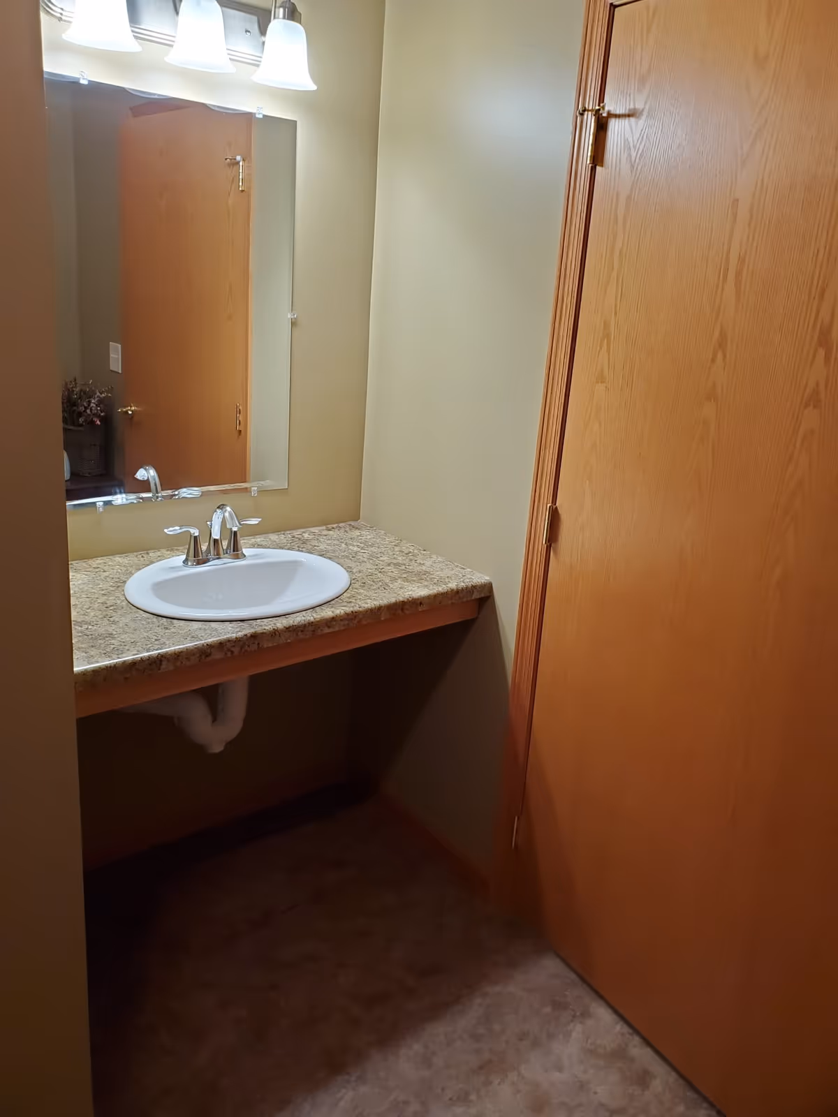 Small bathroom area with a wall-mounted mirror above a countertop with a white sink and silver faucet. There is a wooden door to the right and beige walls and flooring.