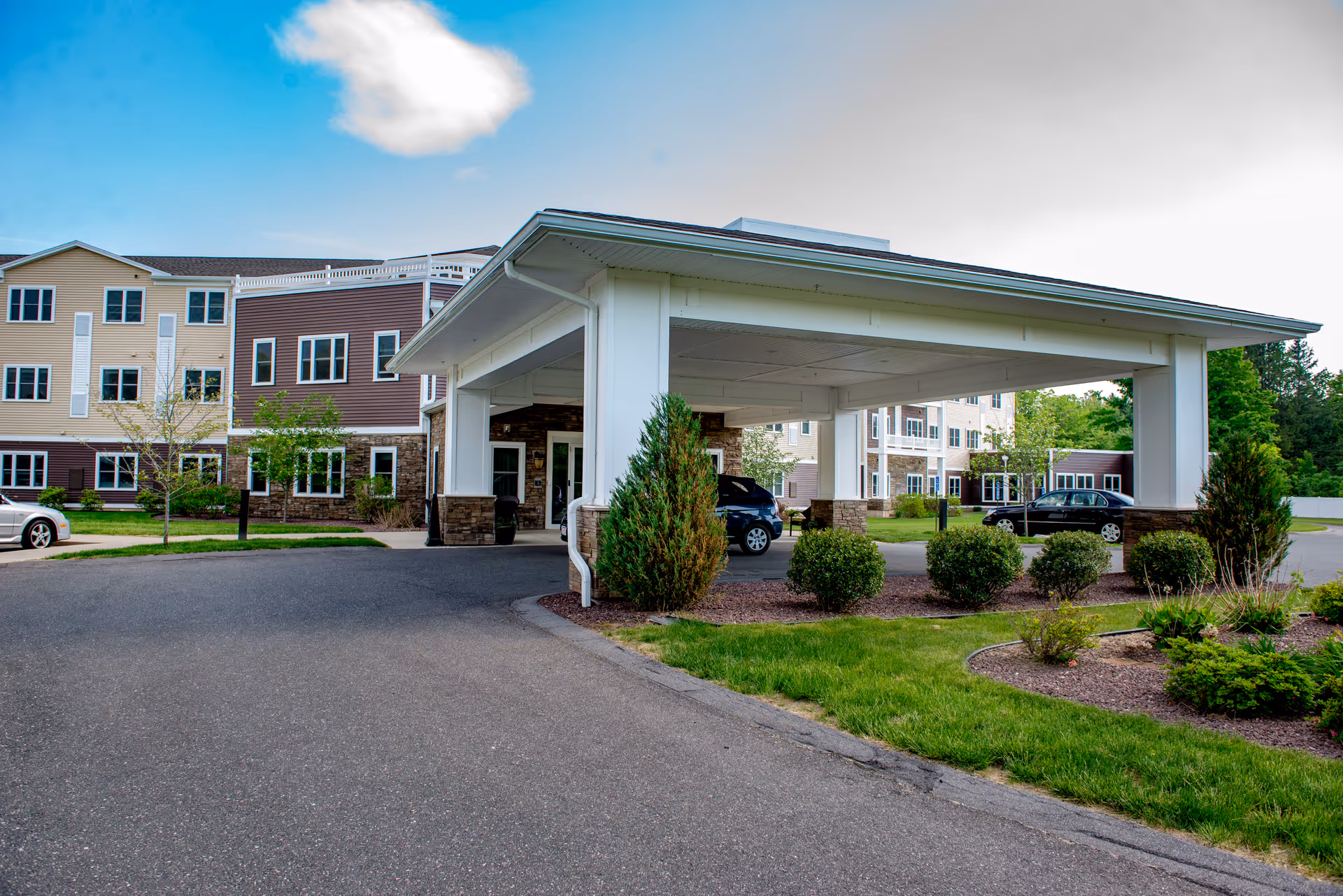Covered porte-cochère entrance of a multi-story assisted living building with landscaping and parked cars.