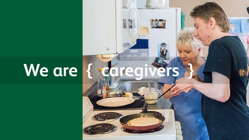 Two caregivers assist an elderly resident at a kitchen stove making pancakes.