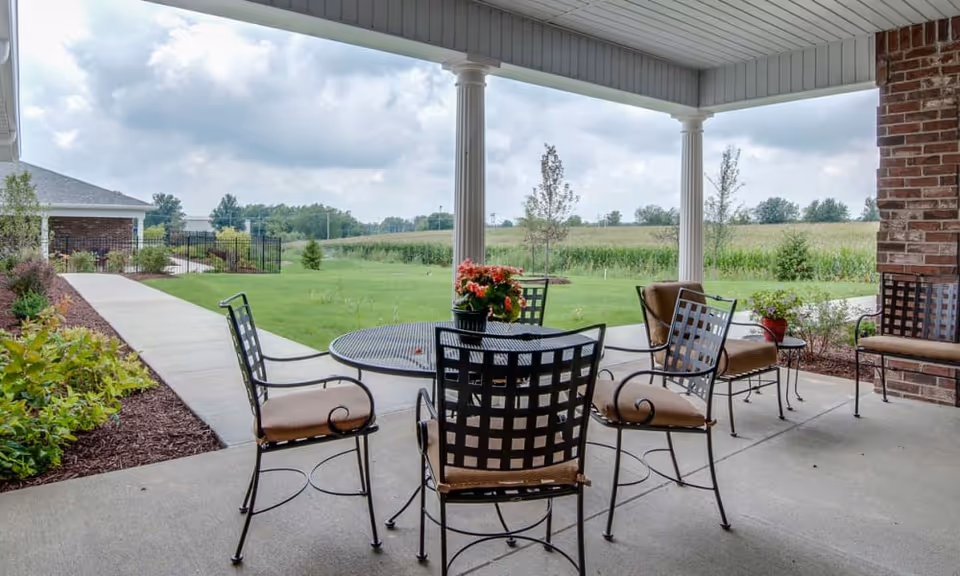 Covered patio with a round metal table and chairs overlooking a grassy lawn and distant field.