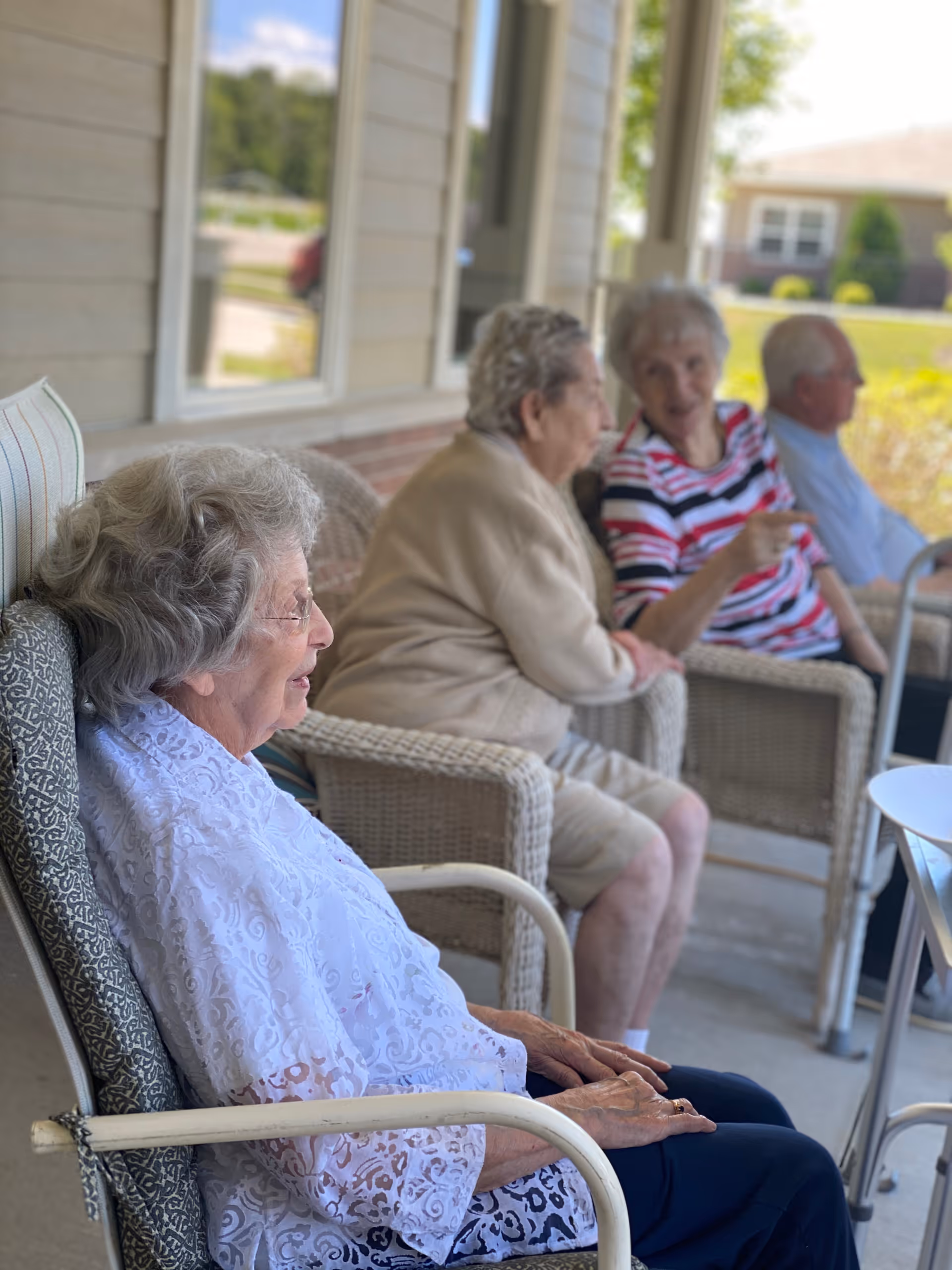 Four elderly individuals sitting on a covered porch in wicker chairs, engaged in conversation and enjoying the outdoor setting at an assisted living facility.