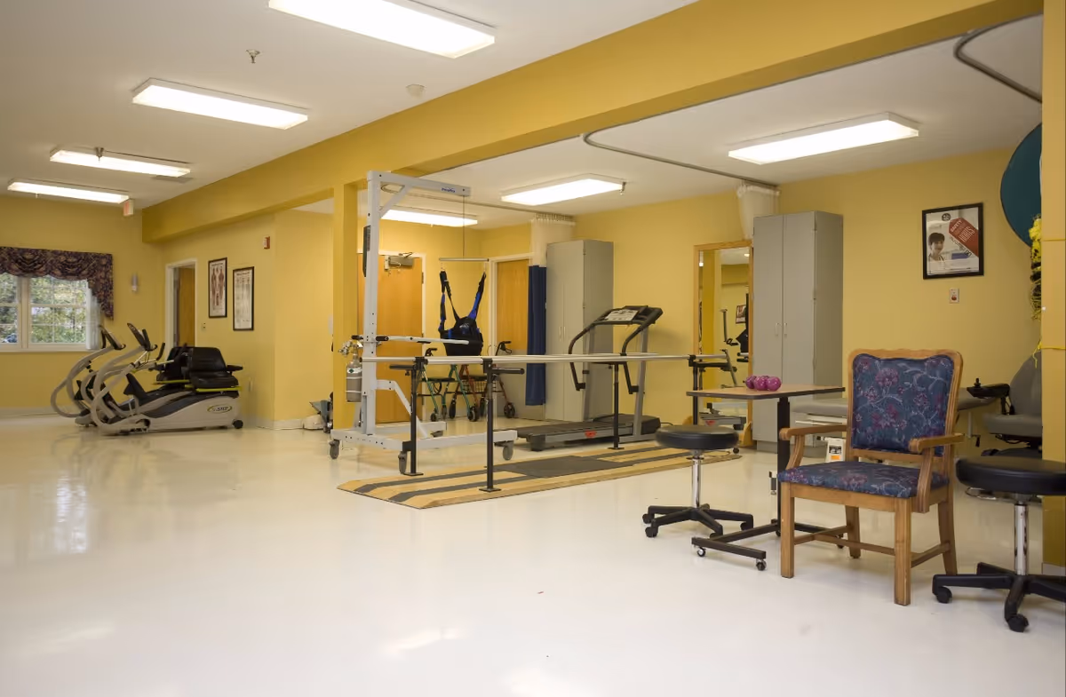A rehabilitation or physical therapy room with exercise equipment including a treadmill, parallel bars, stationary bikes, and a patient lift. The room has yellow walls, white floors, and several chairs and stools. There are cabinets and framed pictures on the walls.