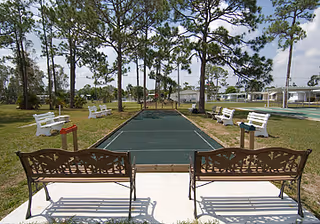 Outdoor recreational area with a bocce ball court surrounded by white benches and two decorative metal benches in the foreground, set among tall pine trees under a partly cloudy sky.