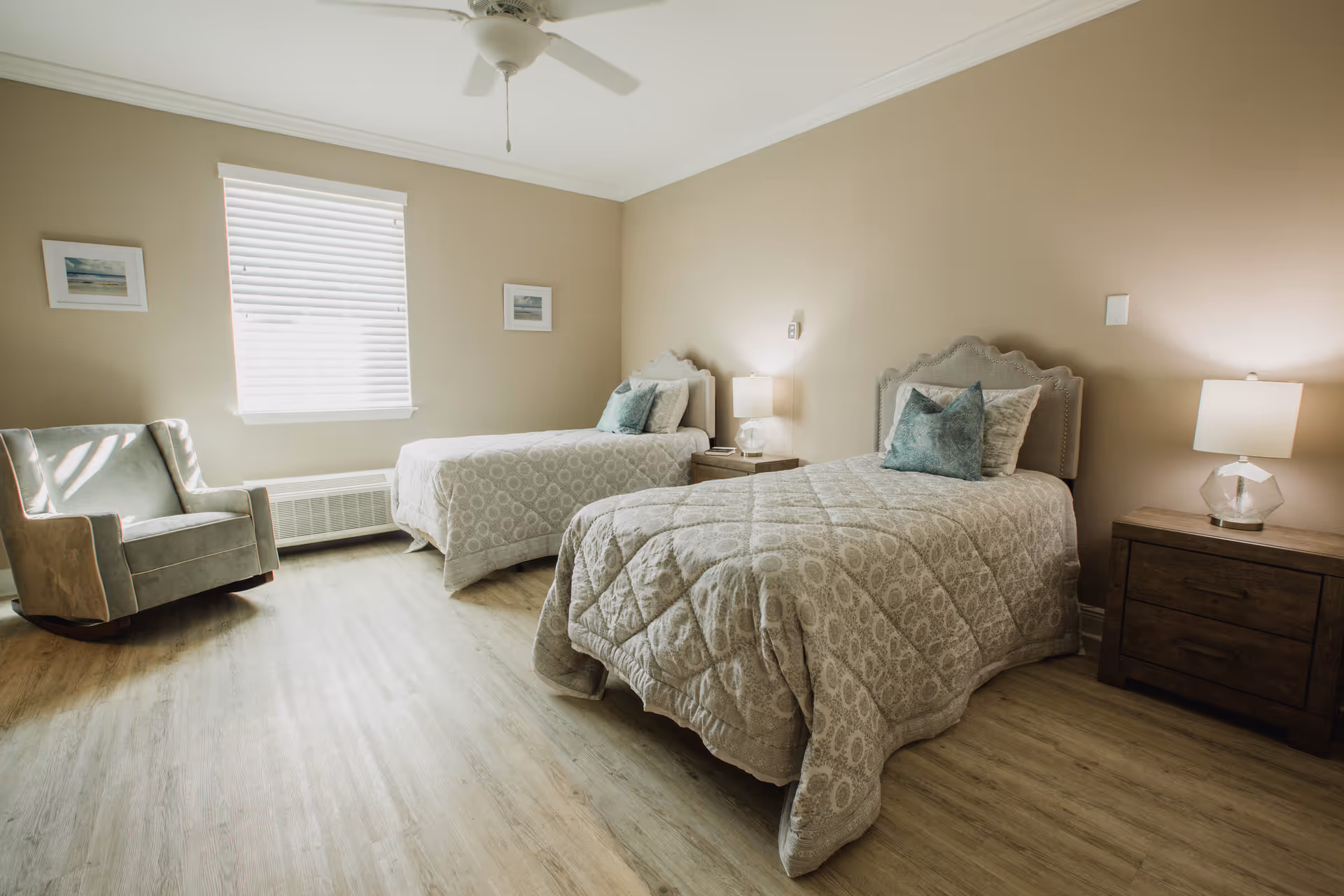 A bright bedroom with two twin beds featuring light-colored patterned bedspreads and teal accent pillows. Each bed has a wooden nightstand with a lamp. There is a comfortable armchair near a window with white blinds, and a ceiling fan above. The walls are painted beige and the floor has light wood-style flooring.