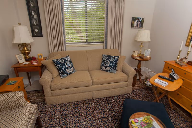 A cozy living room with a beige sofa adorned with two patterned blue and white pillows. There is a window with blinds and curtains behind the sofa. On either side of the sofa are wooden side tables with lamps, framed photos, and small decorative items. A patterned rug covers the floor, and there is a small wooden folding table with a remote control and papers on it. A cushioned ottoman with magazines is also visible in the foreground.