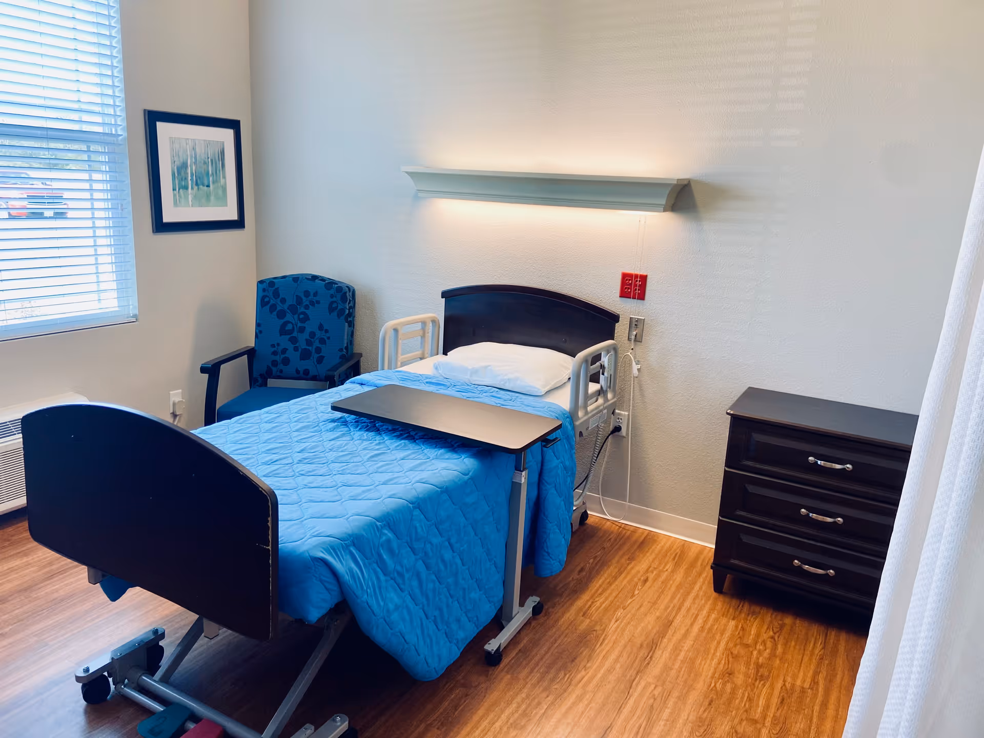 Single patient bedroom with blue quilted bed, over bed tray, chairs, and dresser.