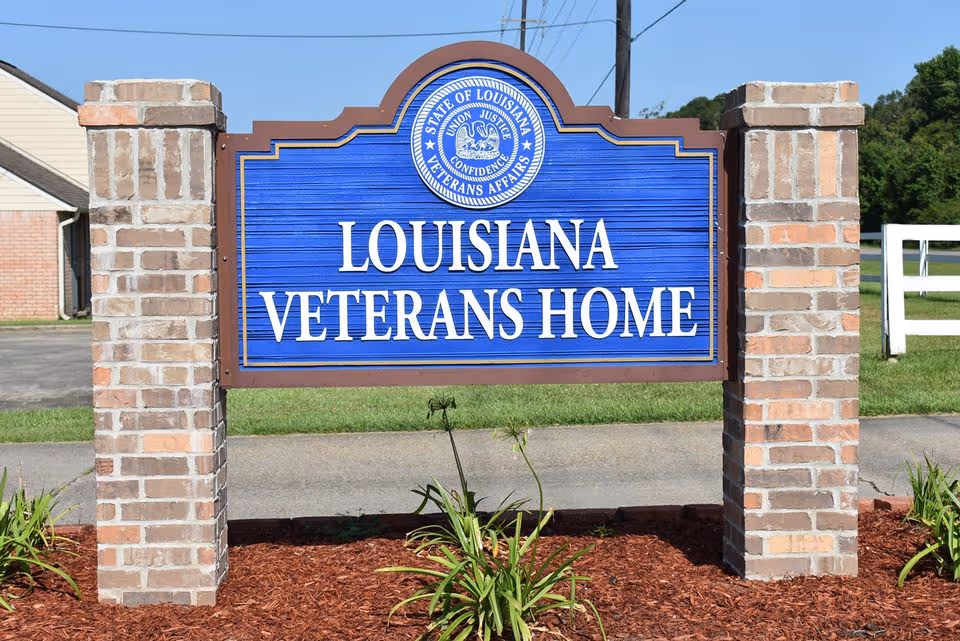 A blue Louisiana Veterans Home sign is framed by brick pillars in a front yard.