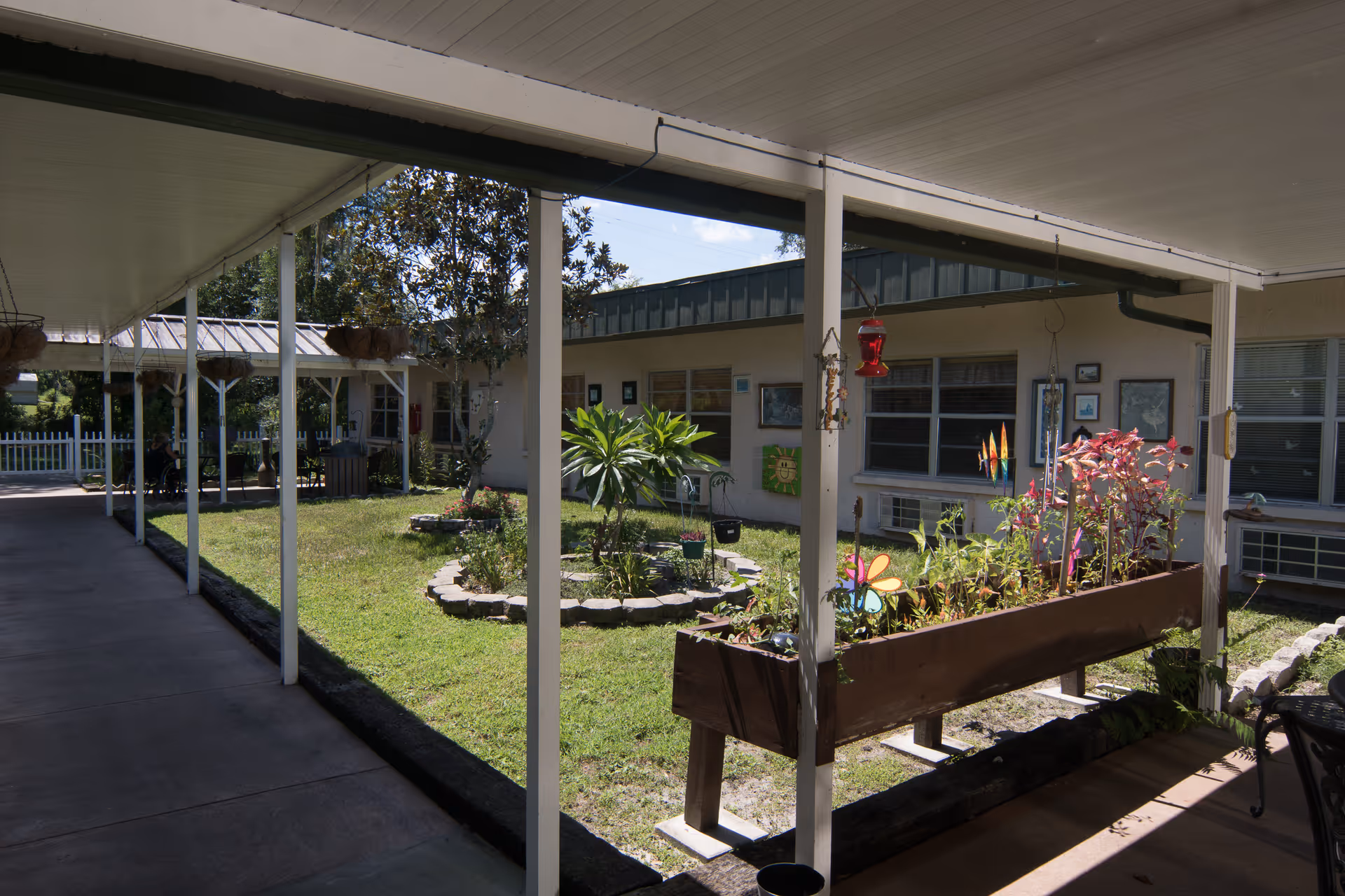 Covered outdoor walkway surrounding a small garden area with plants and decorative items at Royal Oak Nursing Center. The garden has a circular stone border and a raised planter box with colorful plants and wind spinners. The building windows and air conditioning units are visible in the background.