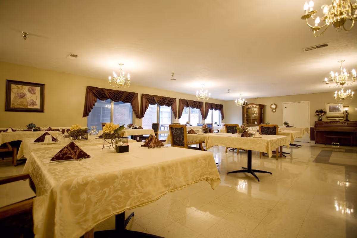 A spacious dining room with multiple tables covered in cream-colored tablecloths, each set with folded napkins and small floral centerpieces. The room features large windows with dark brown valances, chandeliers hanging from the ceiling, and a piano against the far wall.