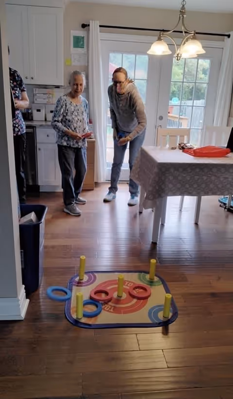 Two elderly women playing a ring toss game inside a room with wooden flooring. One woman is holding a ring, preparing to toss it, while the other watches. The room has a table with a patterned tablecloth, white chairs, and a glass door leading outside. A hanging light fixture is above the table.