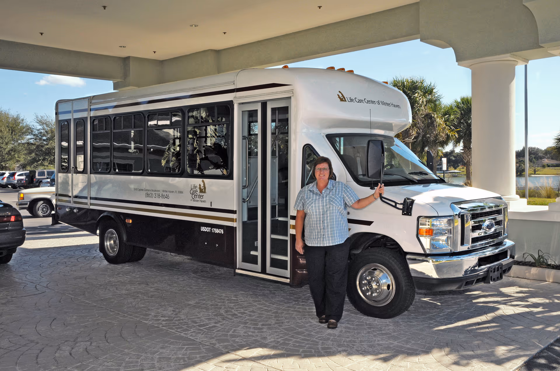A woman standing next to a white and black shuttle bus with the logo and name 'Life Care Center of Winter Haven' parked under a covered driveway with columns and a paved surface.