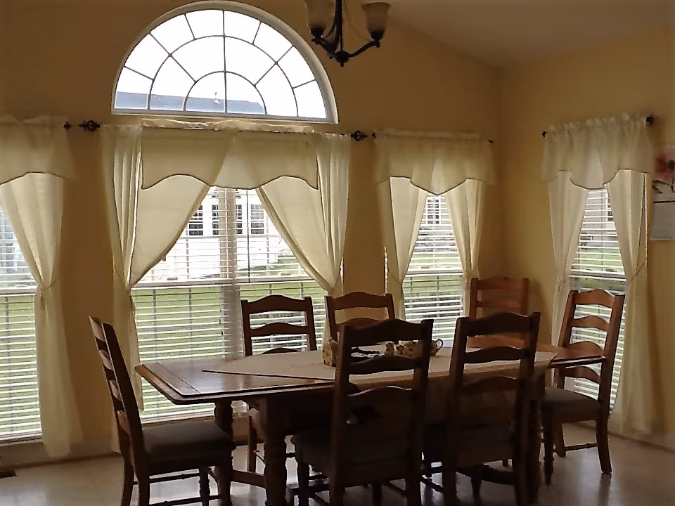 Dining room with a wooden table and chairs set in front of large arched and rectangular windows dressed with cream curtains.