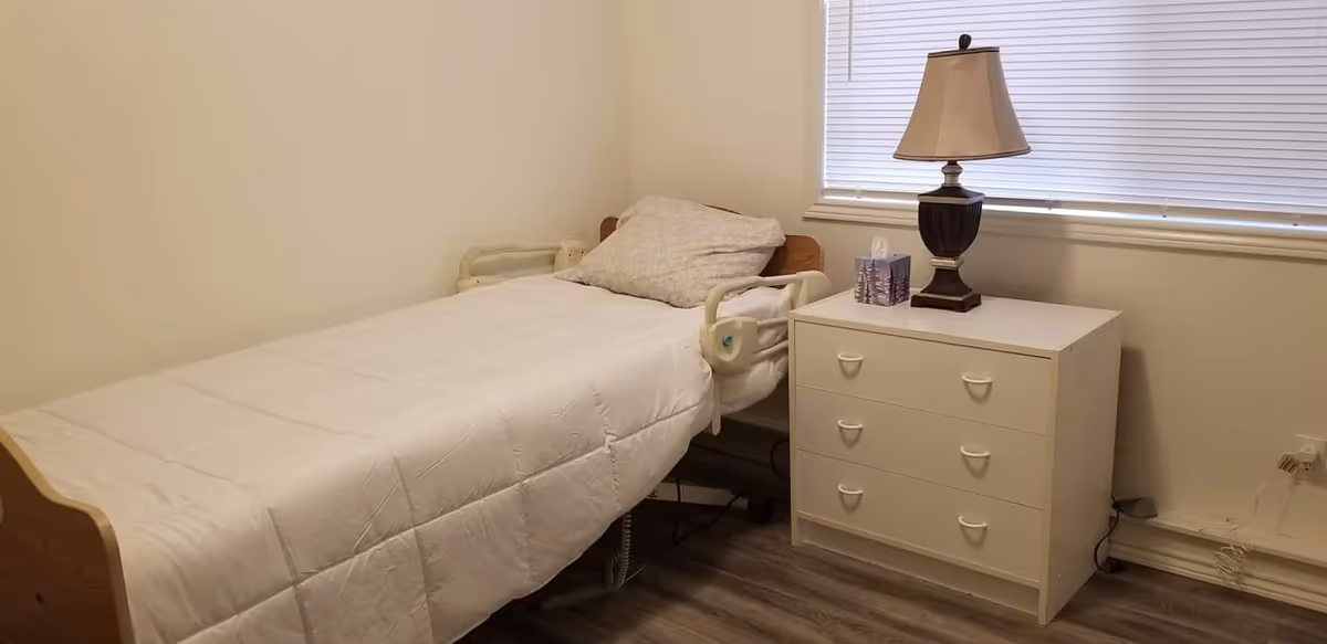 Single hospital-style bed with side rails, white bedding, beside a nightstand and dresser with lamp.
