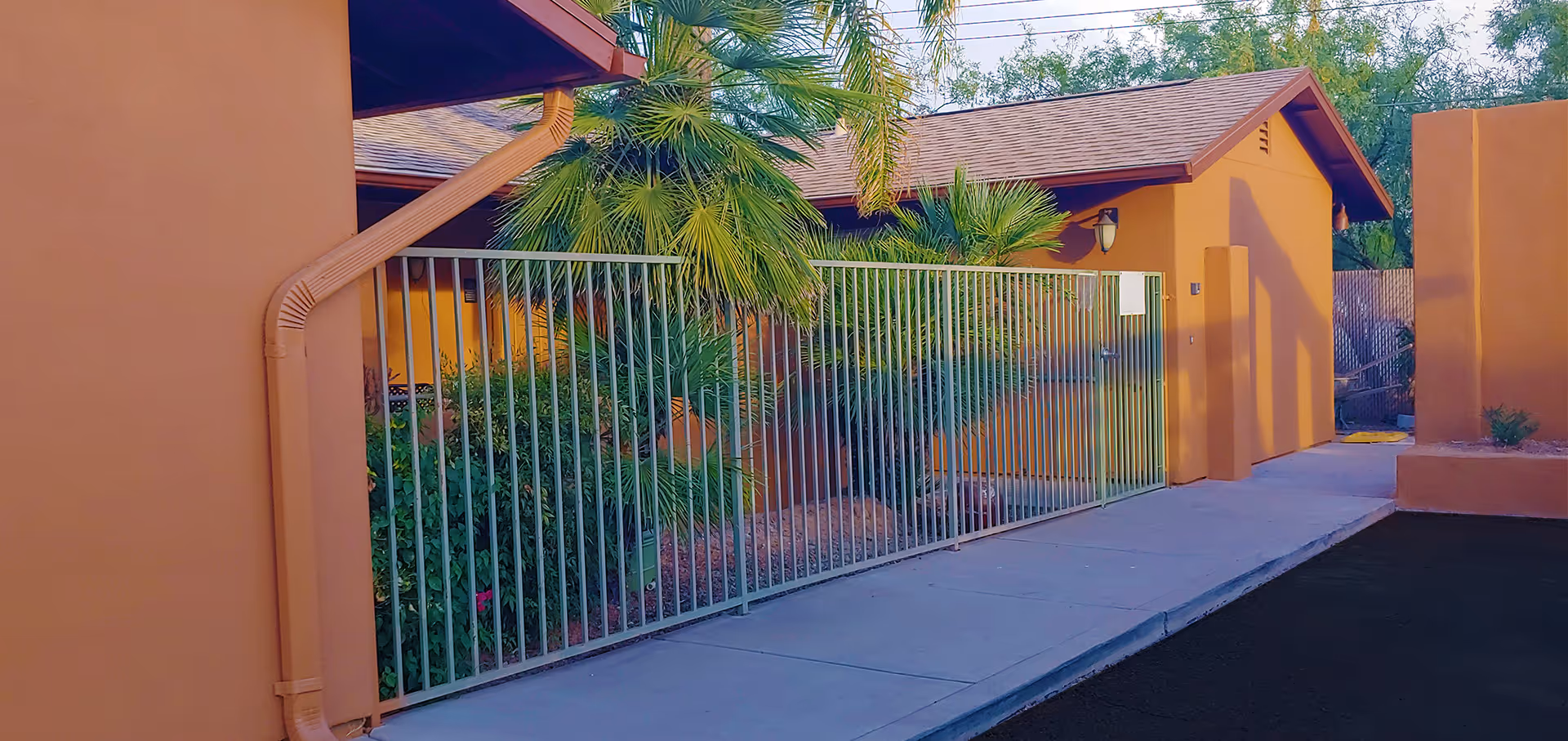 Exterior view of a building with orange walls and a brown roof, featuring a metal fence and gate in front of a landscaped area with palm trees and bushes. A sidewalk runs alongside the building.