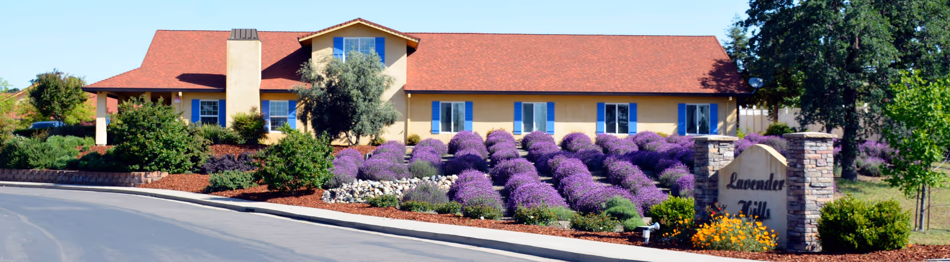 Single-story assisted living facility with purple shrubs, a stone sign, and red-tiled roof.