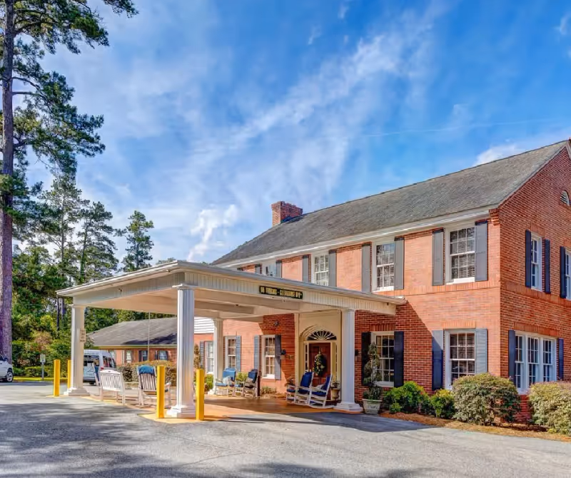 Front entrance of a two-story brick building with a covered porte-cochère, rocking chairs on the porch, and surrounding landscaping.