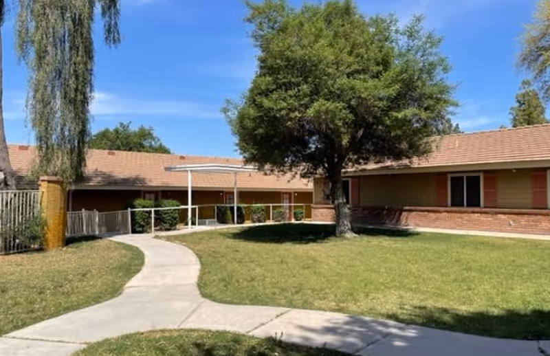 Single-story brick senior living facility exterior with curved walkway, lawn, and trees.