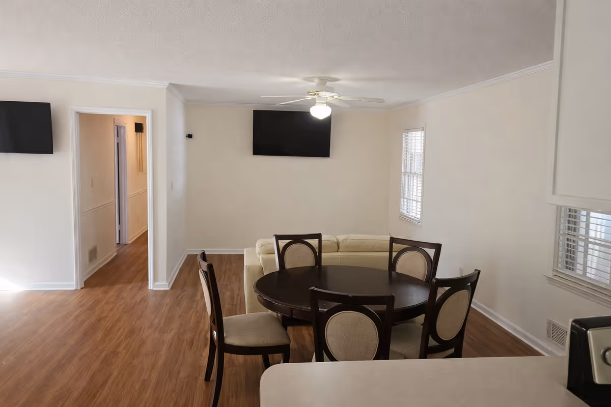 Bright dining area with round table and six chairs, sofa, TVs, and hallway doorway.