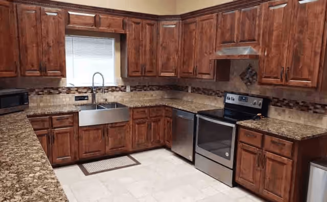 A kitchen with wooden cabinets, granite countertops, a stainless steel double sink under a window, a stainless steel dishwasher, and a stainless steel electric stove with an oven. The backsplash features a mosaic tile design, and the floor is tiled in a light color.