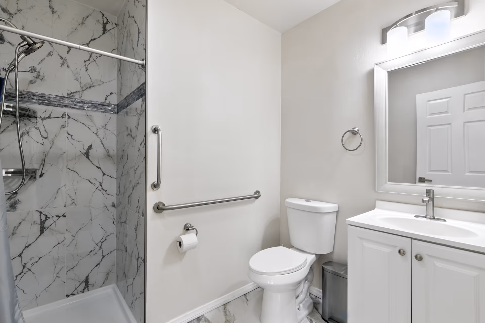 Clean bathroom with marble shower, grab bars, toilet, and white vanity in a senior living facility.