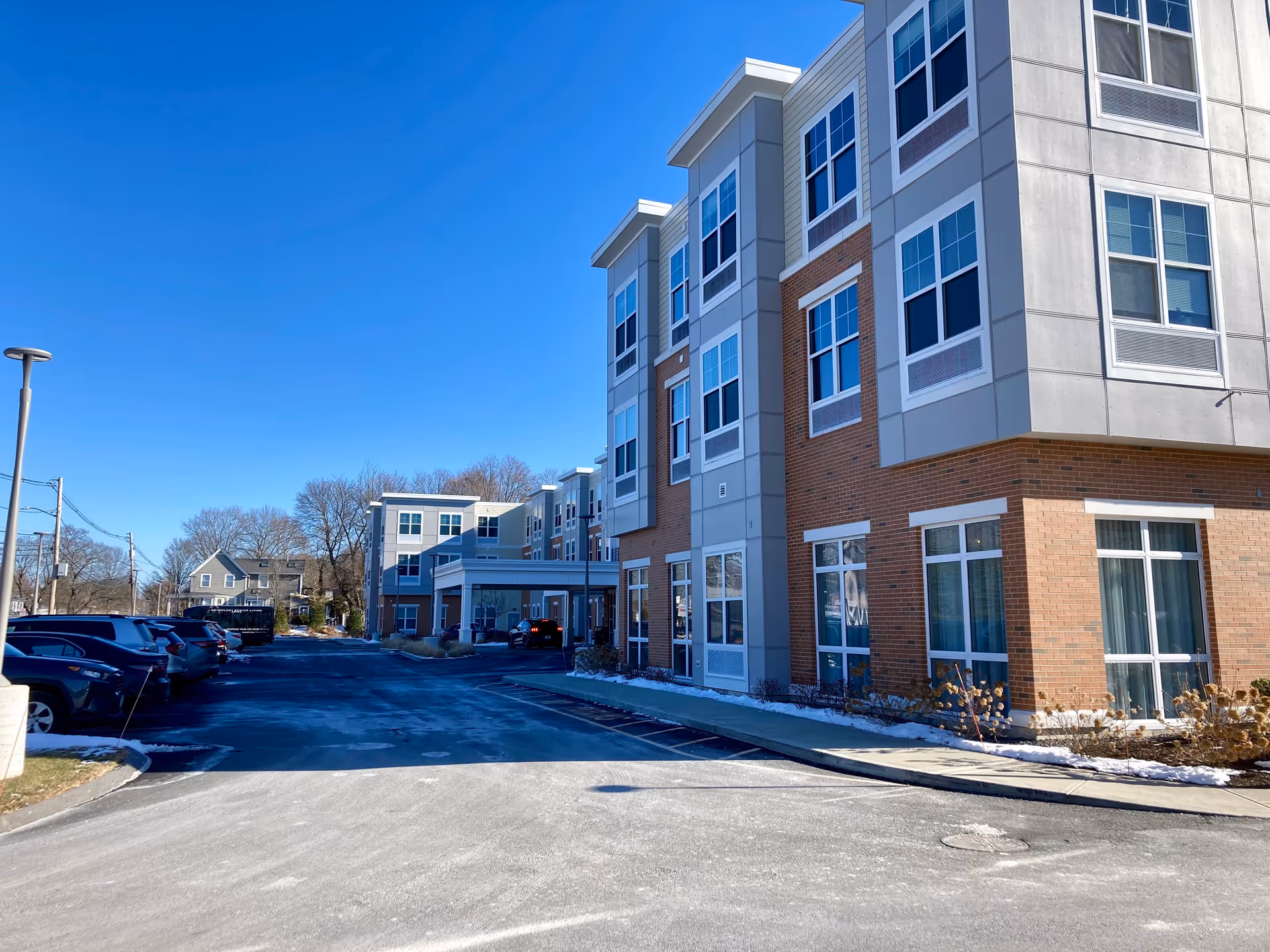 Exterior view of a modern three-story senior living facility building with large windows and a covered entrance. Several cars are parked along the driveway, and there is a clear blue sky above.