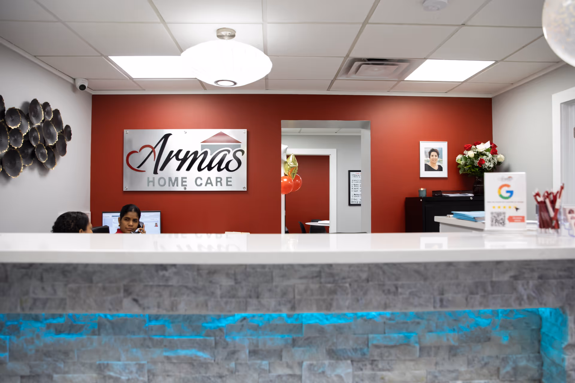 Reception area with Armas Home Care sign behind a marble-front desk and two staff members.