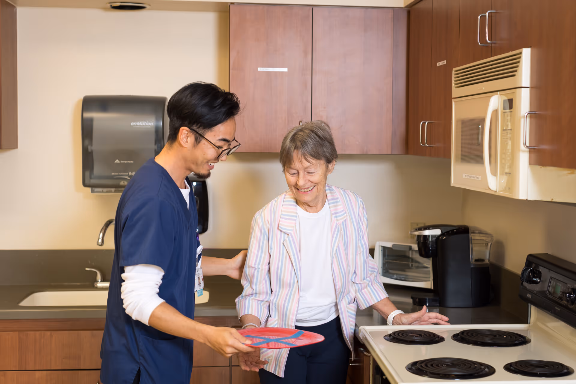 A young male caregiver in blue scrubs assists an elderly woman in a striped jacket in a kitchen area. The woman is smiling and standing near a stove while the caregiver holds a red plate. The kitchen has wooden cabinets, a microwave, a coffee maker, and a sink.