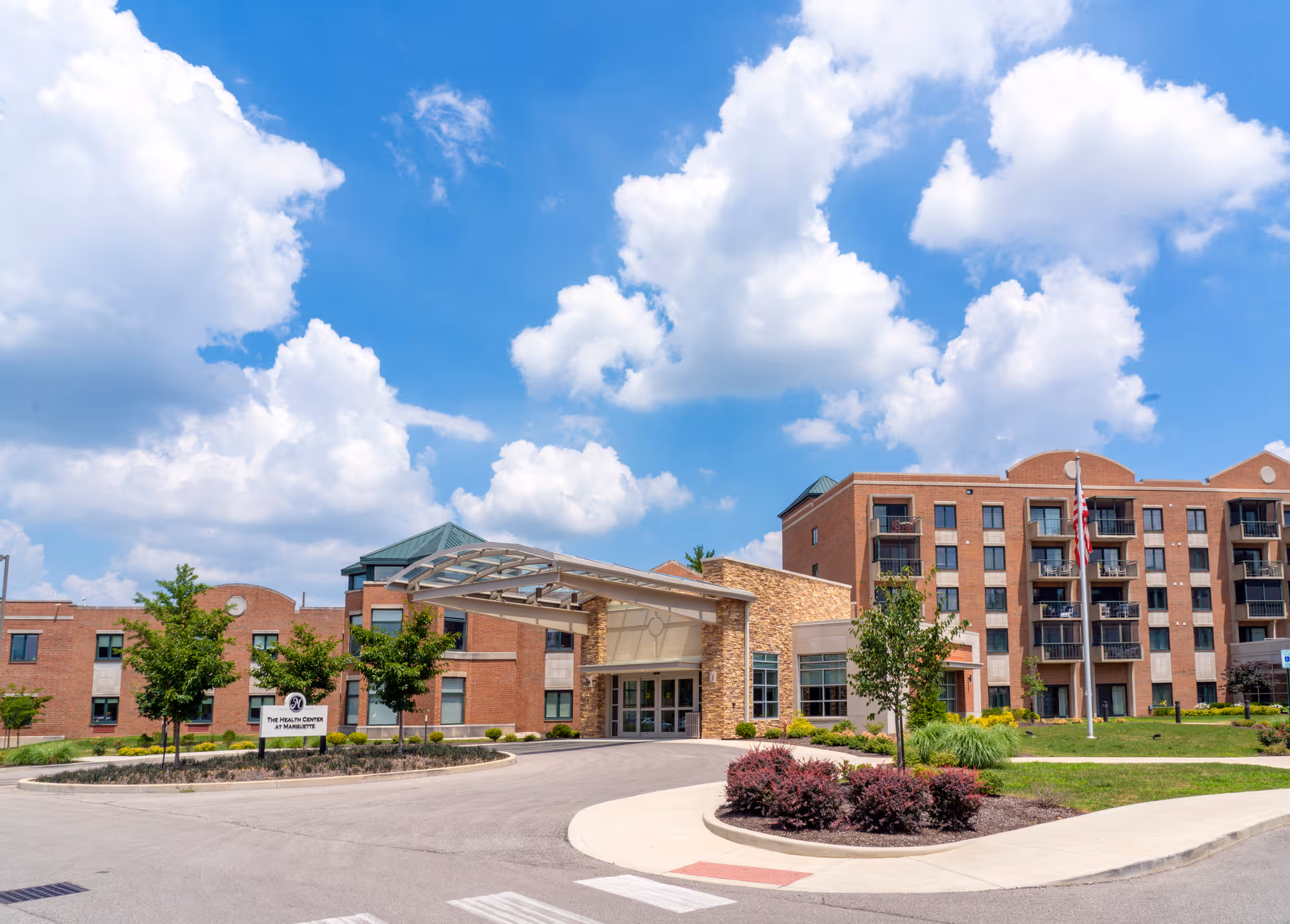 Exterior view of a brick senior living facility entrance with landscaped grounds and blue sky.