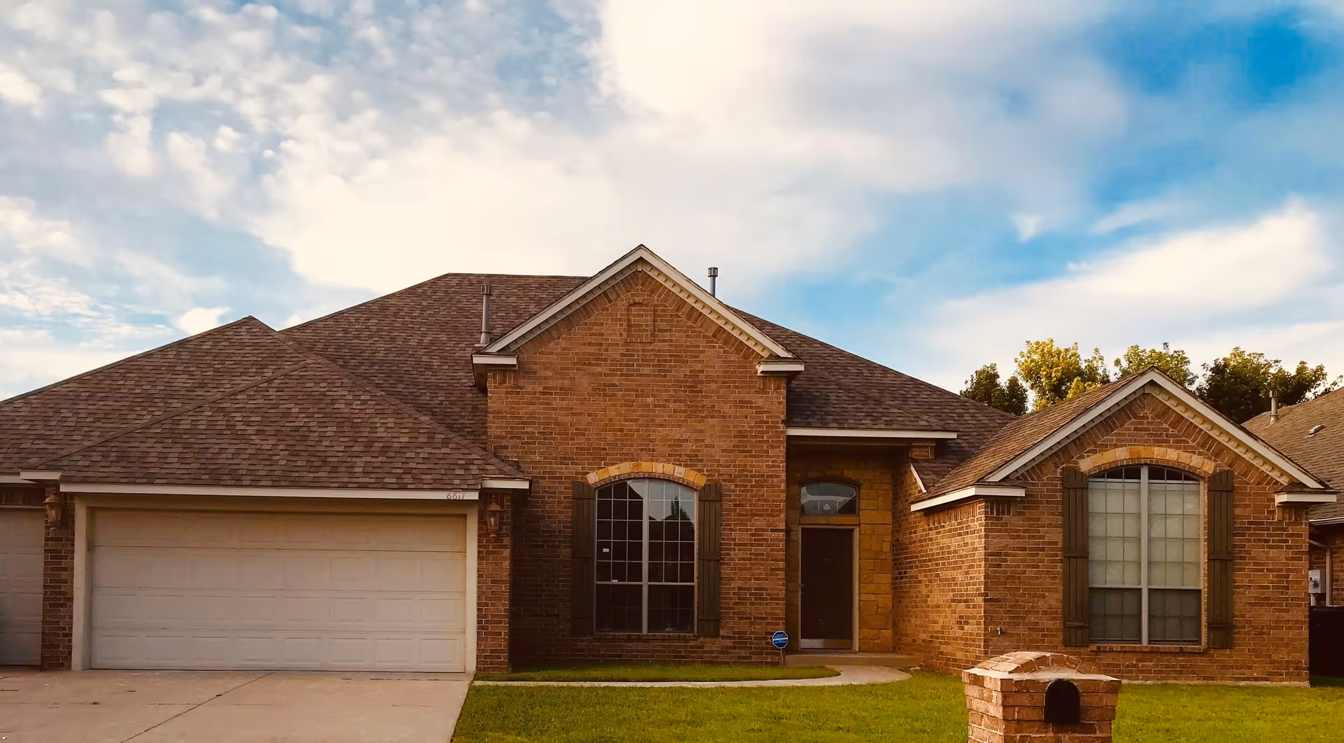 Front exterior of a single-story brick suburban house with a garage, lawn, and blue sky.