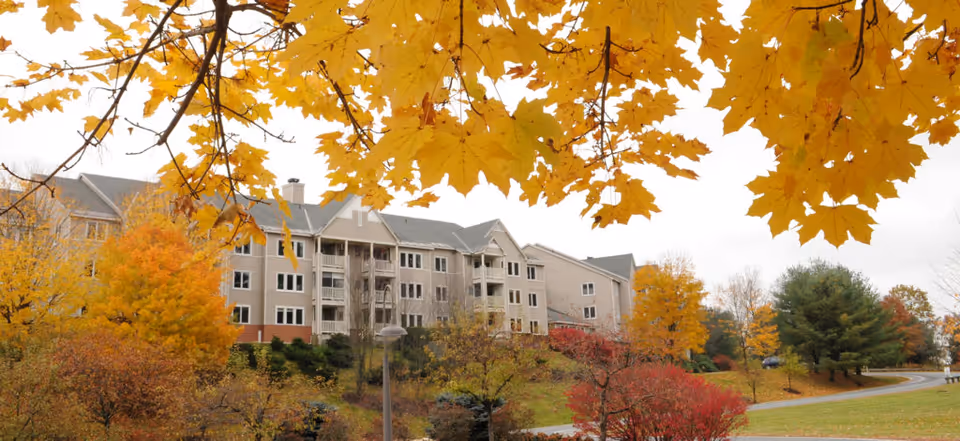 Exterior view of a multi-story senior living facility building surrounded by trees with autumn foliage in shades of yellow, orange, and red. A curved driveway and green lawn are visible in the foreground under an overcast sky.