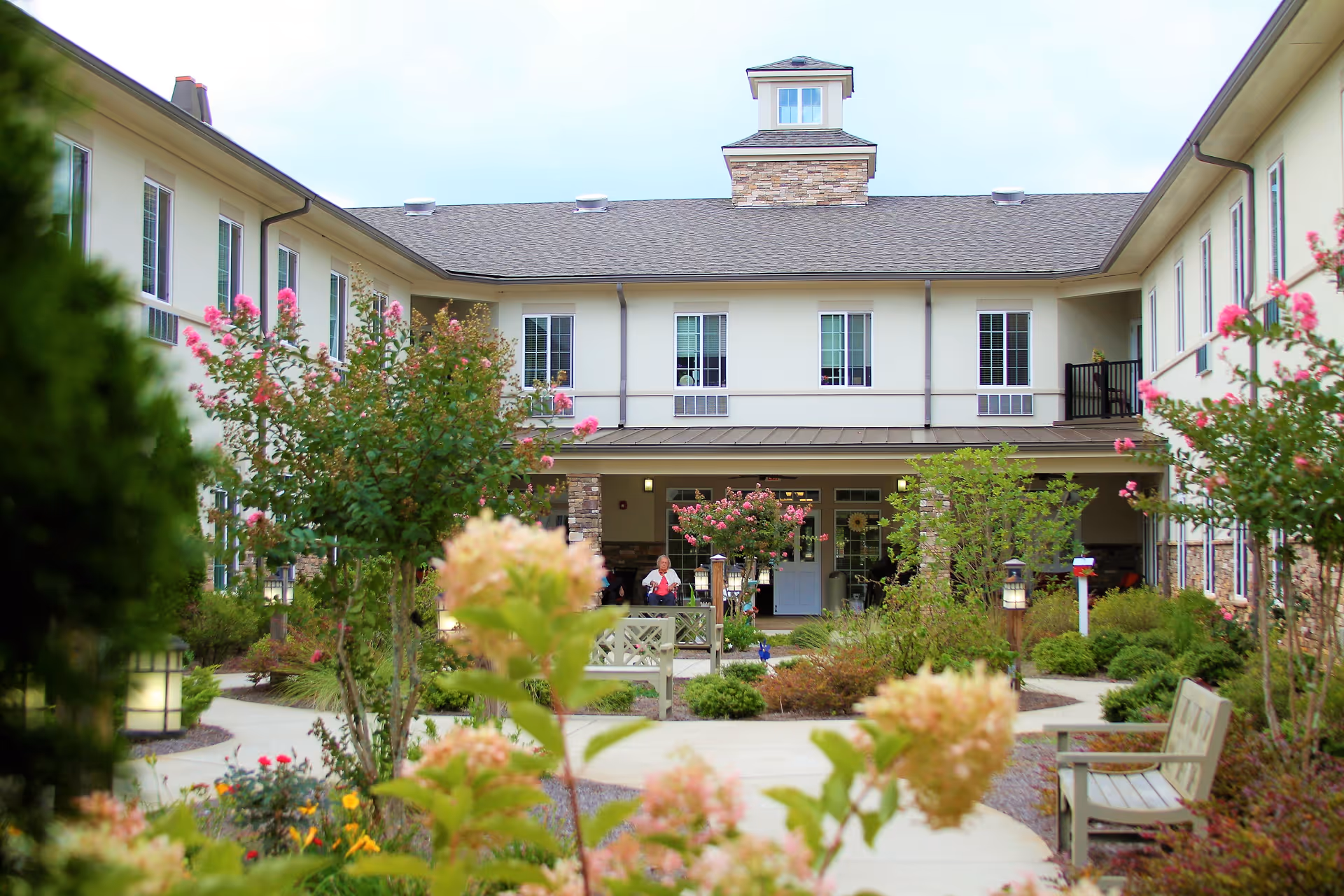 Outdoor courtyard area of The Landings of Canton Hills senior living facility with a paved walkway, benches, flowering bushes, and a two-story building with multiple windows in the background.