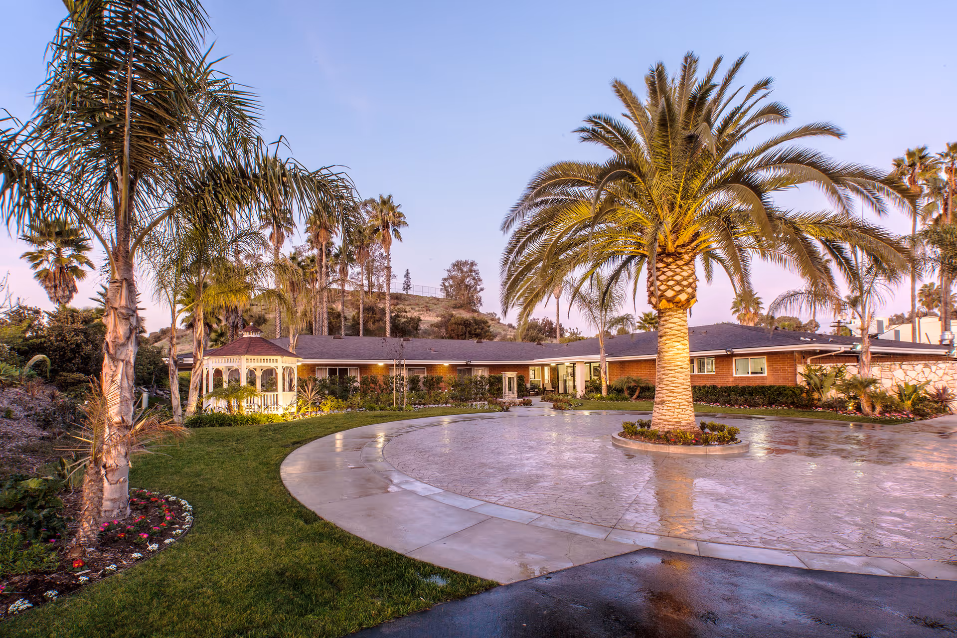 Exterior view of University Care Center featuring a circular driveway with a large palm tree in the center, surrounded by well-maintained landscaping including grass, flower beds, and additional palm trees. The building is a single-story structure with a gazebo on the left side and a backdrop of hills and a clear sky at dusk.