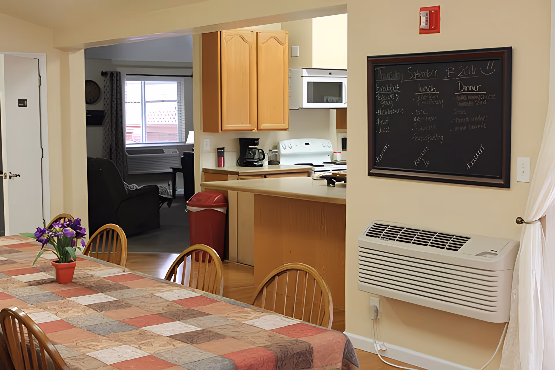 Interior view of a senior living facility showing a dining area with a table covered by a multicolored tablecloth and wooden chairs. A small potted plant with purple flowers is on the table. In the background, there is a kitchen with wooden cabinets, a white stove, microwave, and coffee maker. A red trash bin is near the kitchen counter. A blackboard with handwritten notes is mounted on the wall above a white wall-mounted heater. A living room area with a recliner and window is visible beyond the kitchen.