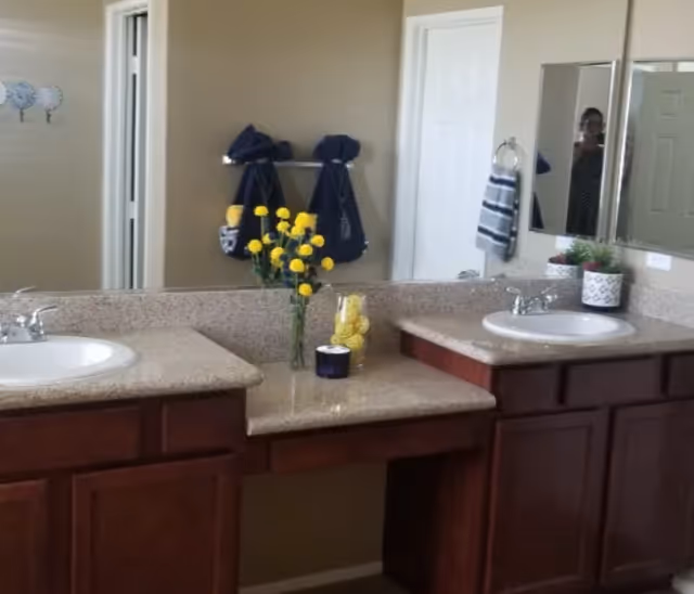 Dual-sink bathroom vanity with beige countertops, dark wood cabinets, and a vase of yellow flowers.