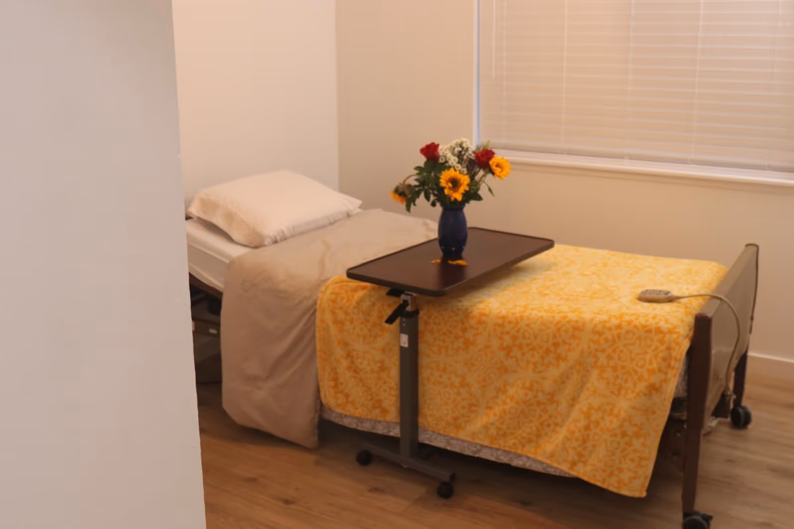 A small resident bedroom with a bed, yellow blanket, flowers, and window blinds.
