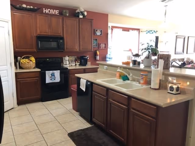 A kitchen area with wooden cabinets, a black stove and microwave, a double sink, and a countertop with various items including a basket of bananas, a coffee maker, and a candle. The kitchen opens into a bright living space with windows and some furniture visible in the background.