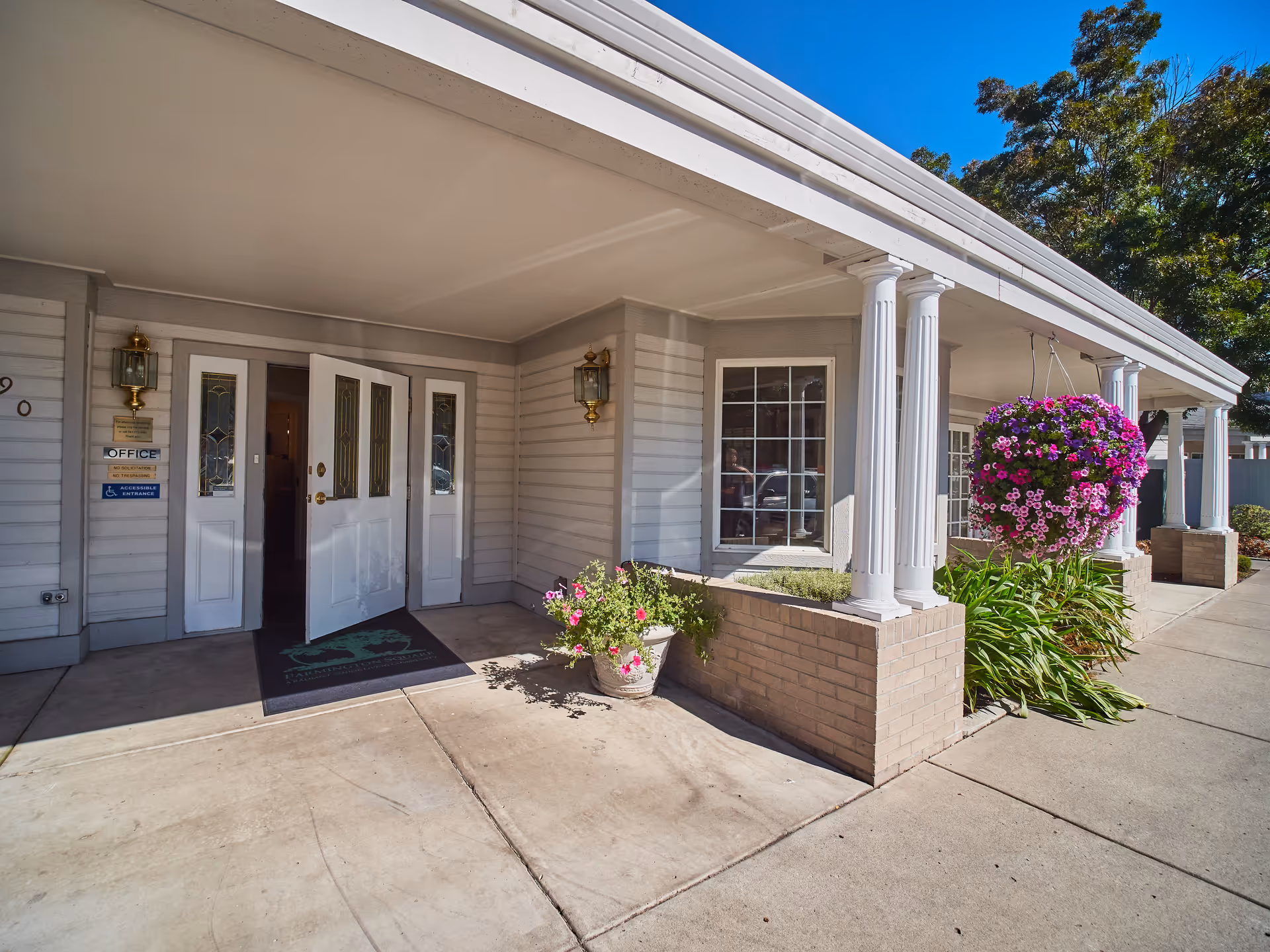 Covered front entrance of a senior living facility with white columns, open double doors, potted flowers and a hanging flower basket.