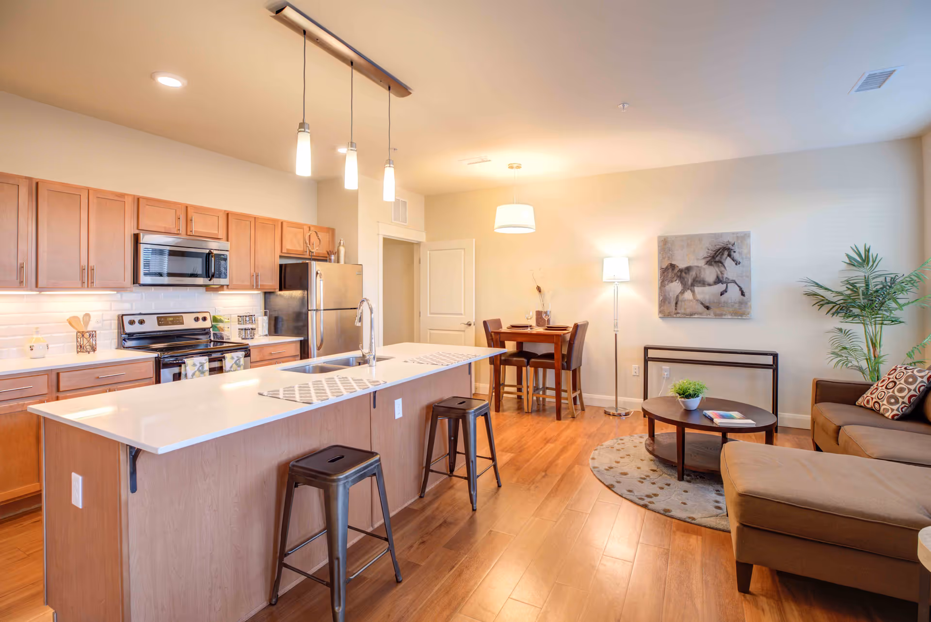 Open-concept interior of an apartment featuring a kitchen with wooden cabinets, stainless steel appliances, and a large island with two metal stools. Adjacent to the kitchen is a small dining area with a wooden table and four chairs, illuminated by a hanging light fixture. The living area includes a brown sectional sofa, a round coffee table with a plant and a book, a floor lamp, a framed horse artwork on the wall, and a potted plant in the corner. The space has wooden flooring and warm lighting.