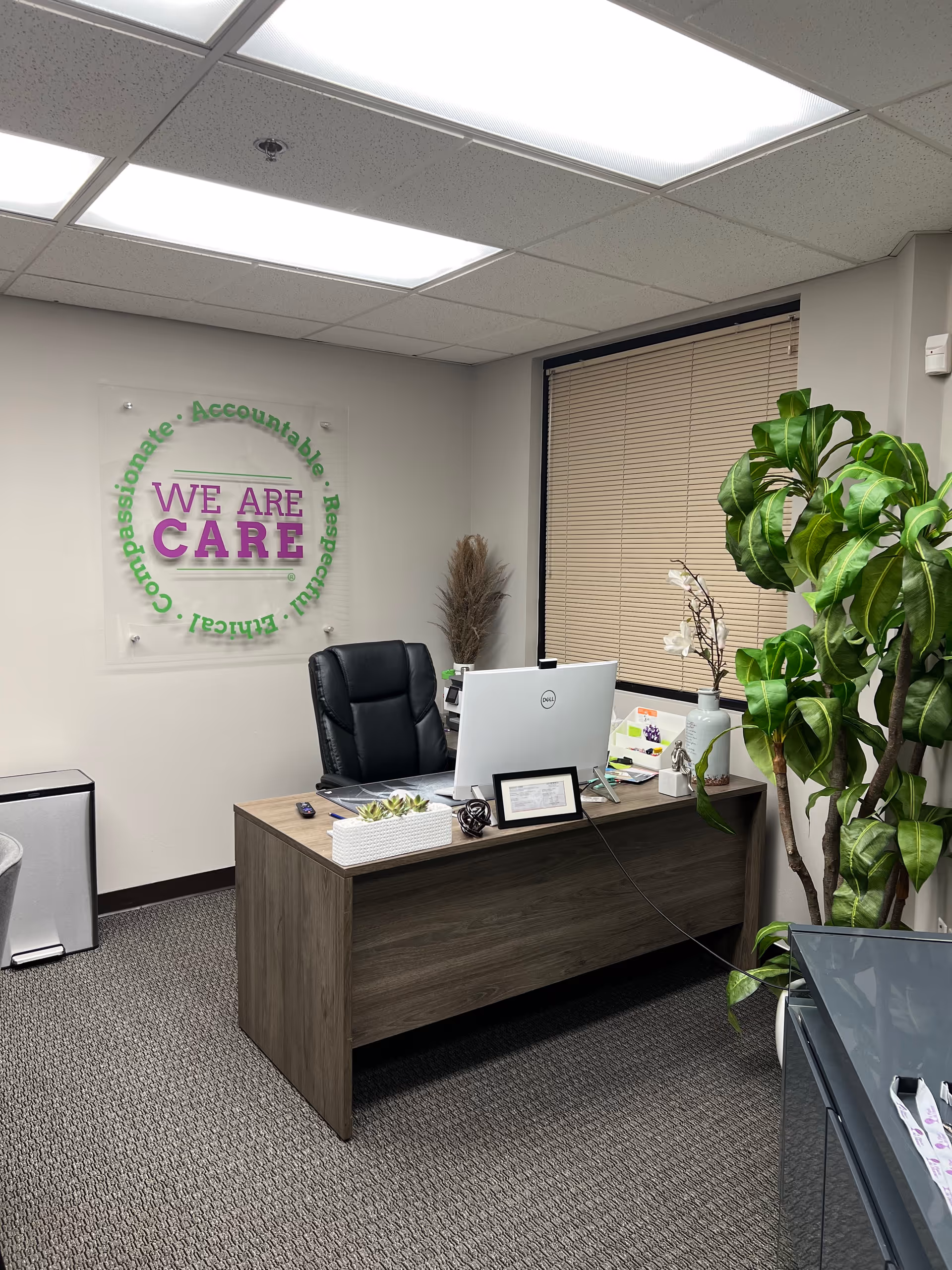 Office desk with computer, sign reading 'We Are Care', plants, and window blinds.