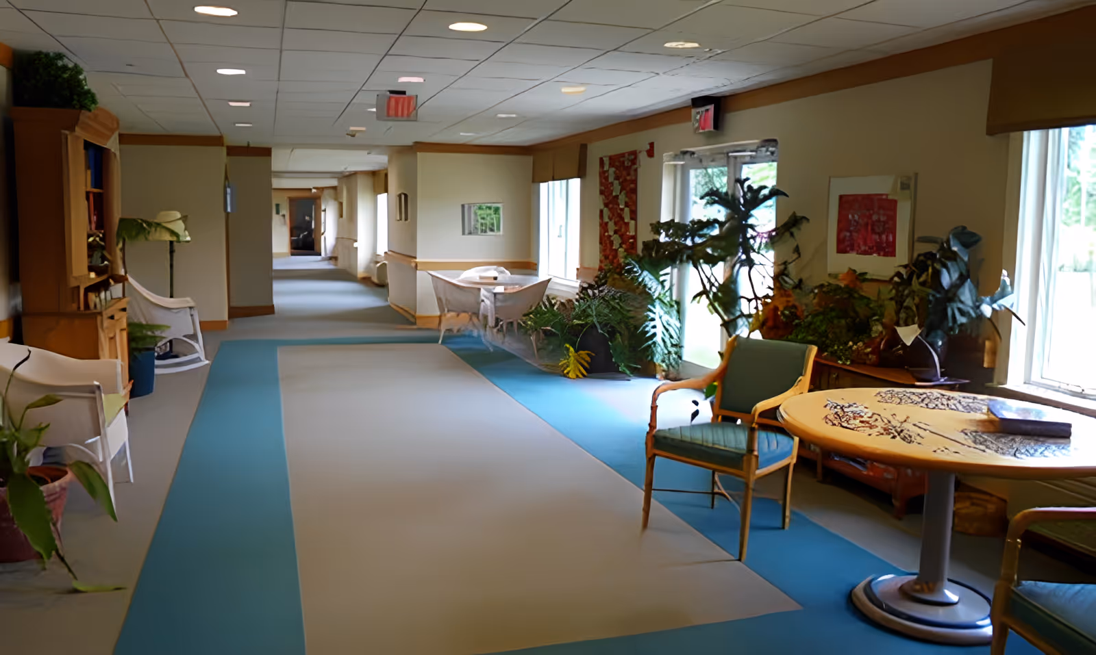 A long, carpeted hallway in an assisted living facility with chairs, tables, plants, and windows along the right side. The hallway is well-lit with ceiling lights and natural light from the windows. There are decorative elements such as framed artwork and a wooden cabinet on the left side.