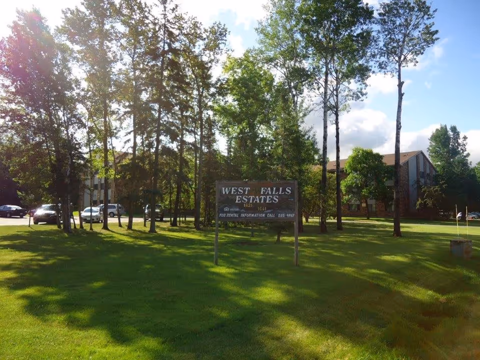 A grassy outdoor area with several tall trees and a sign that reads 'West Falls Estates' along with rental information. In the background, there is a multi-story building partially visible and several parked cars.
