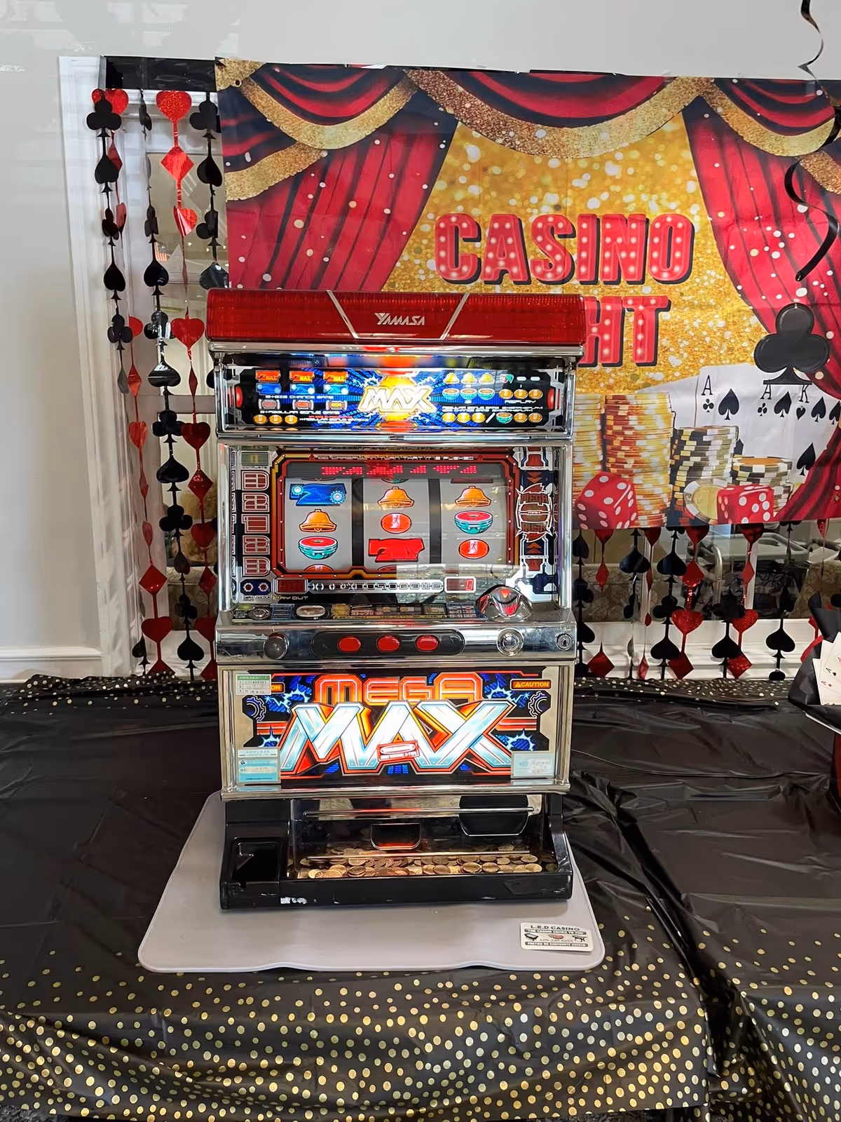 Indoors, a shiny slot machine stands on a decorated table with a casino backdrop.