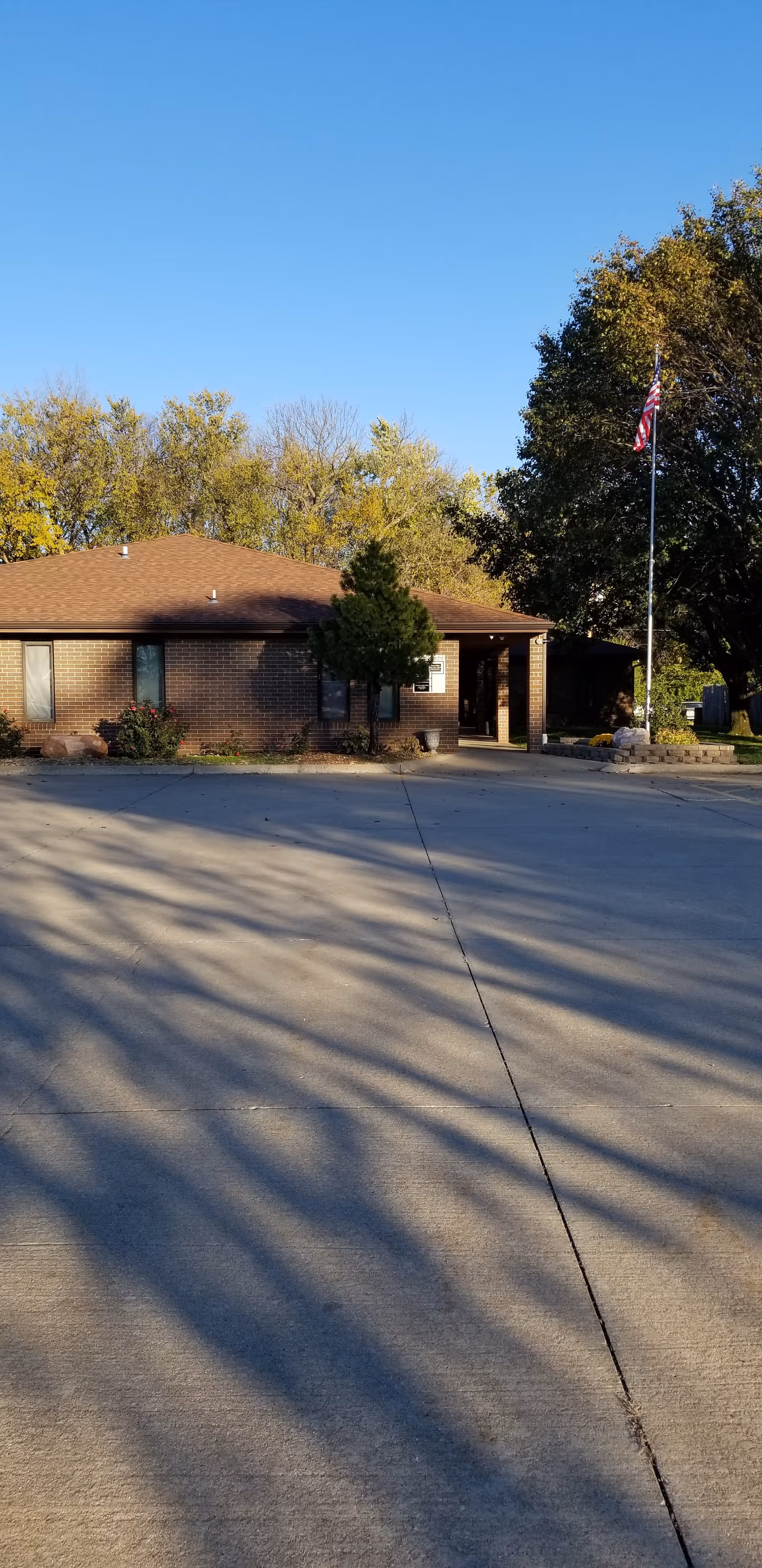 Single-story brick facility entrance with flagpole and trees on a sunny day.