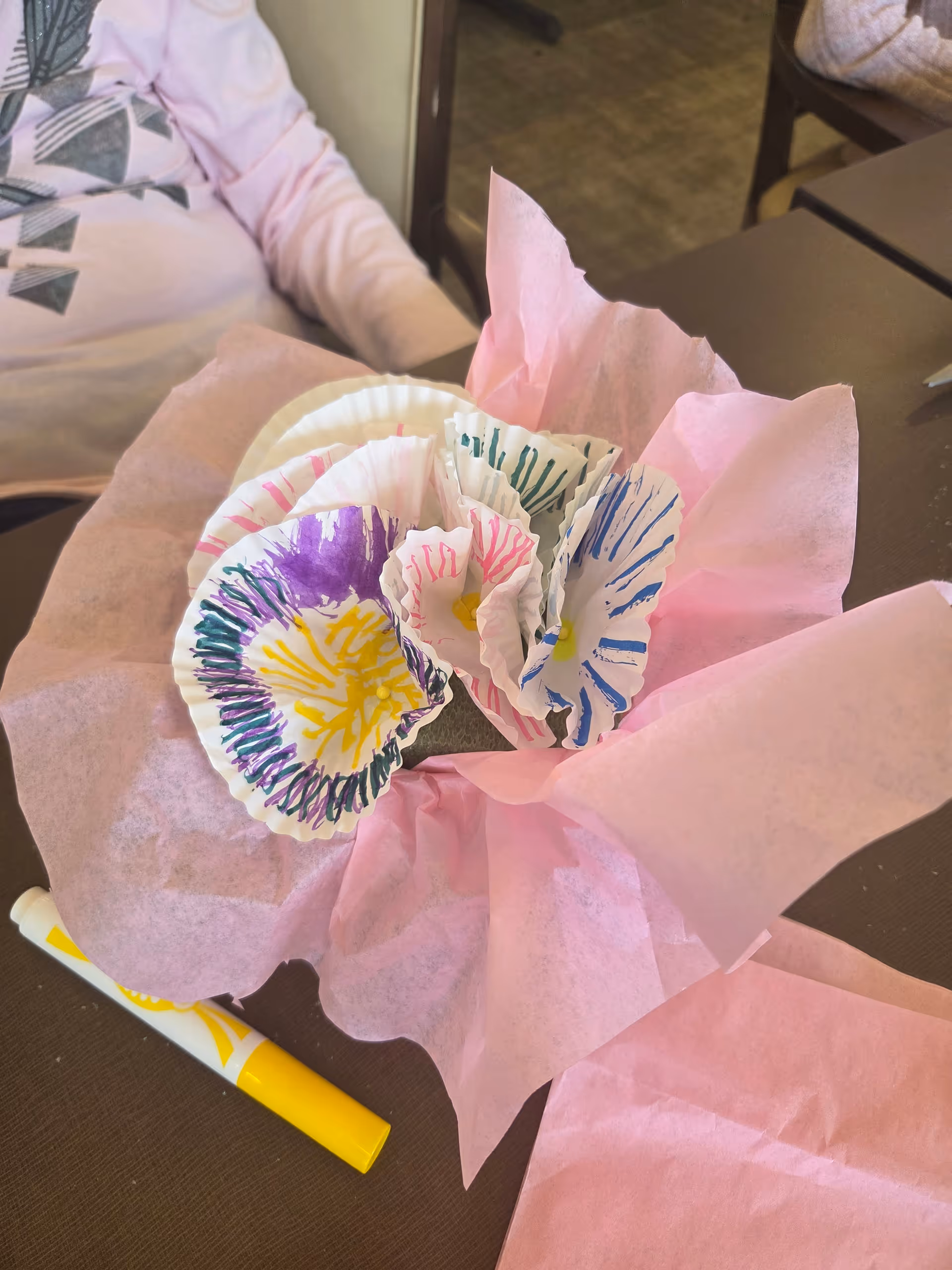 A table with pink tissue paper, colorful handmade paper flowers, and a yellow marker.