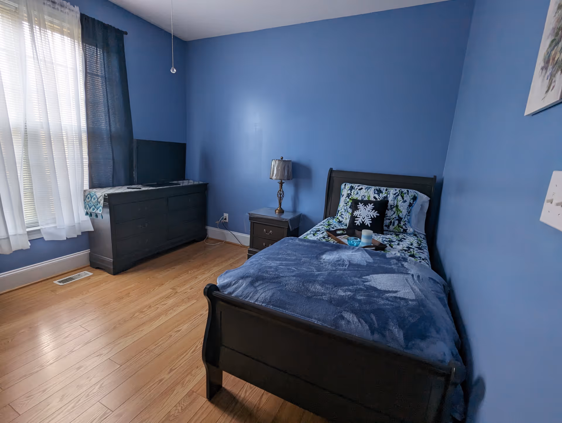 Blue-painted bedroom with a single bed, dresser, nightstand, and window with sheer curtains.