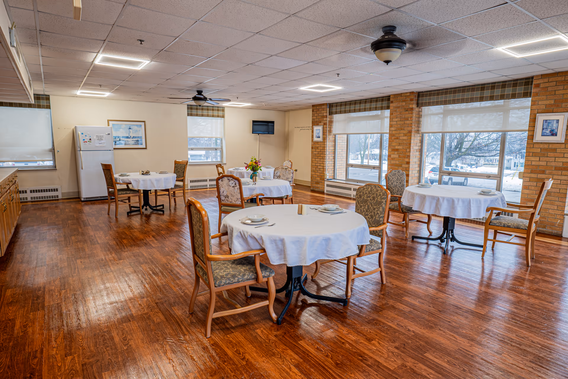A bright dining room with wooden floors and several round tables covered with white tablecloths. Each table is set with plates, bowls, and utensils, and surrounded by cushioned wooden chairs. Large windows with blinds allow natural light to fill the room, and a refrigerator and cabinets are visible along one wall. A small wall-mounted TV is also present.