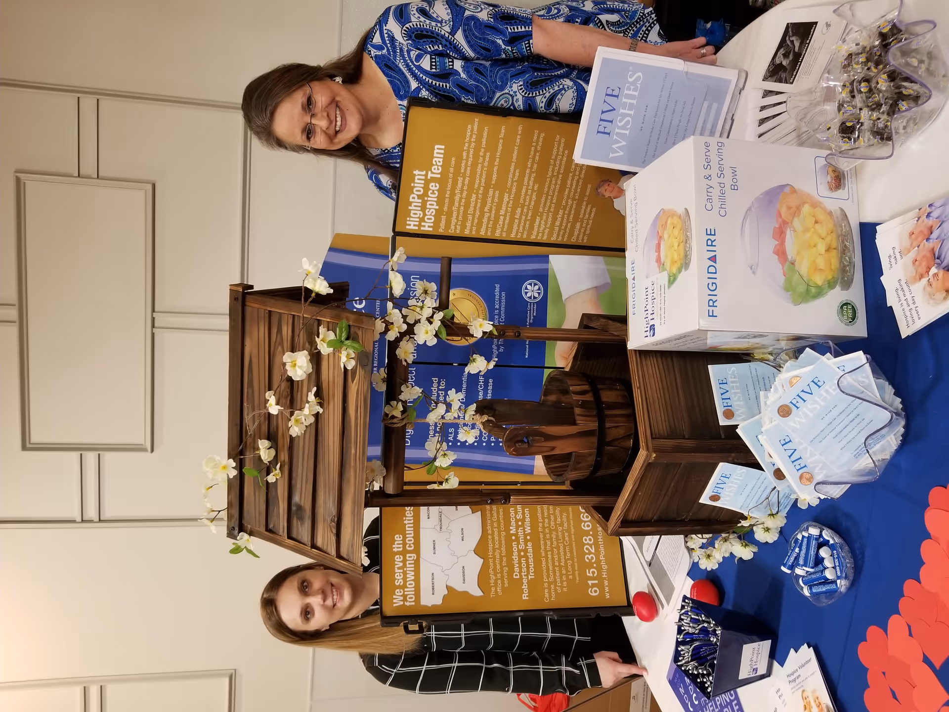 Two smiling women stand beside a HighPoint Hospice informational display with brochures indoors.